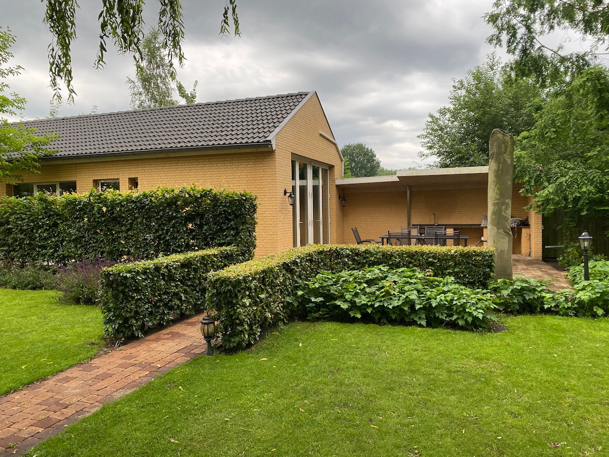 The well-maintained exterior shows a yellow-brick guesthouse bordered by neat hedges and lush greenery. A stone pathway leads to a sheltered terrace with outdoor seating, framed by a soft expanse of grass and flowering plants, all set under an overcast sky.