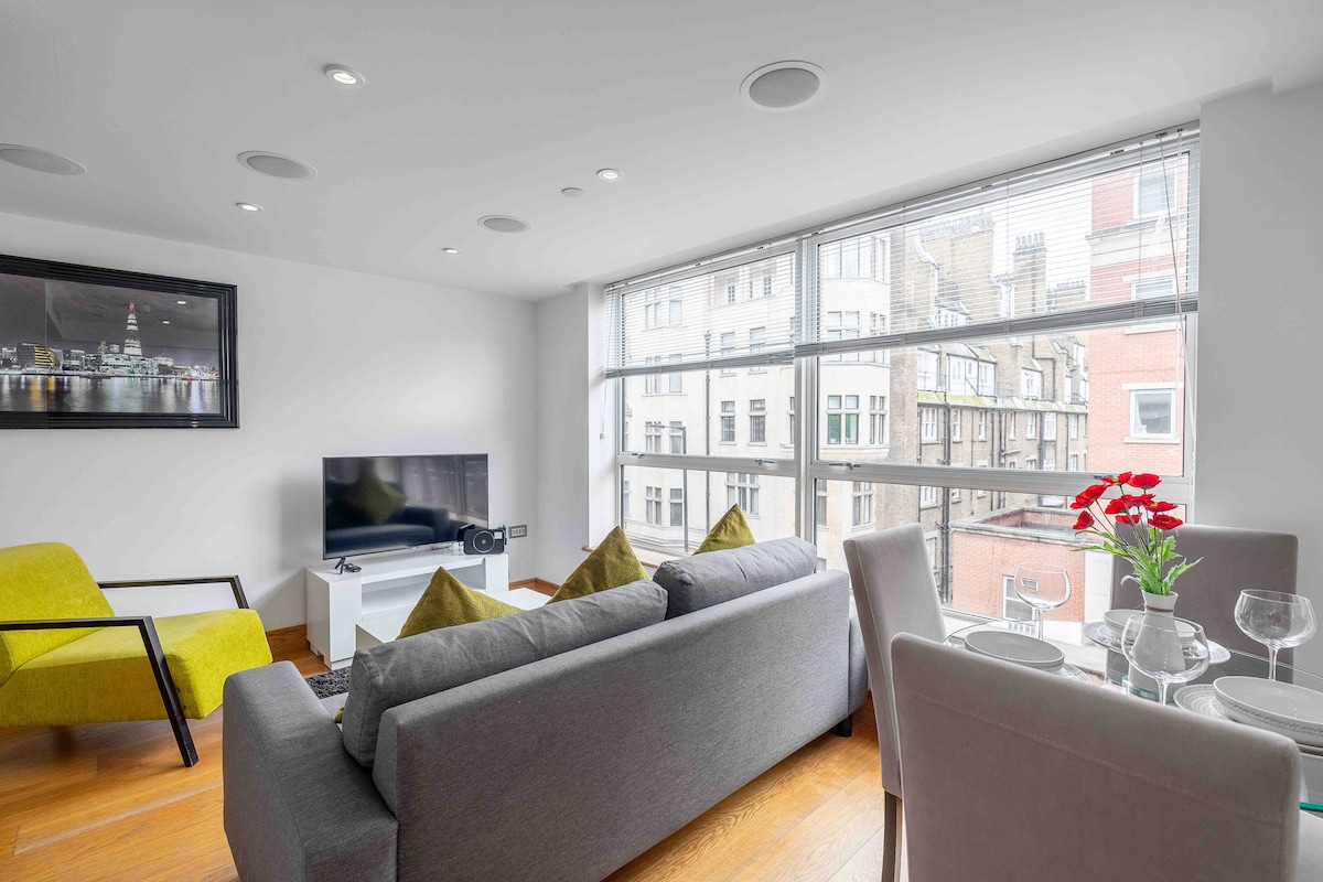 A bright sitting area features large windows providing natural light and views of the surrounding buildings. The space includes a contemporary sofa with decorative cushions, a television mounted on the wall, and a dining table set for four, complemented by an accent chair.