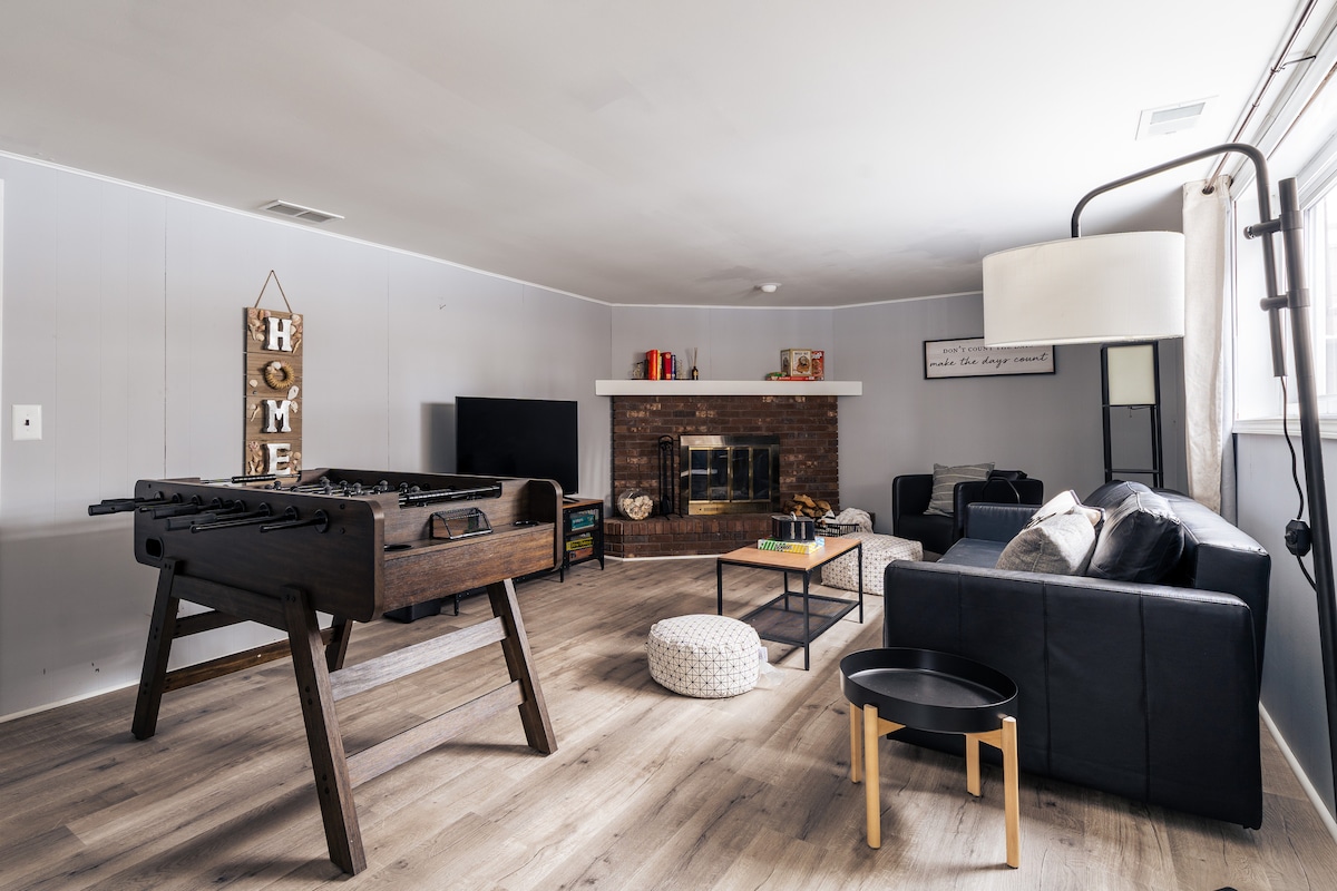 A spacious family room features a foosball table, a fireplace with a brick surround, and a wall-mounted television. Comfortable seating includes a leather sofa and accent chairs, while a light-colored pouf and a small table provide additional decor and functionality.