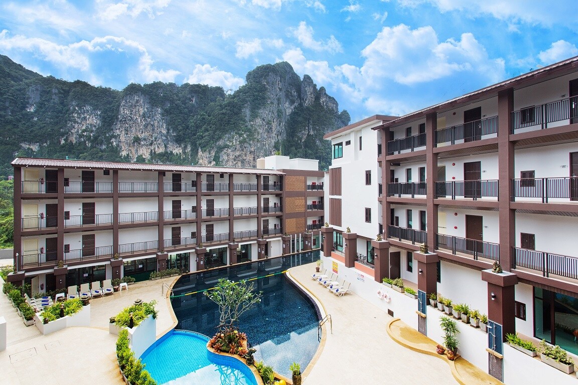 A view of the Lai Thai Luxury Condominiums showcases multiple three-story buildings surrounding a large, curvilinear pool. Lush greenery is present along the pool's edges, and distant limestone cliffs rise against a vibrant sky, contributing to a serene atmosphere.