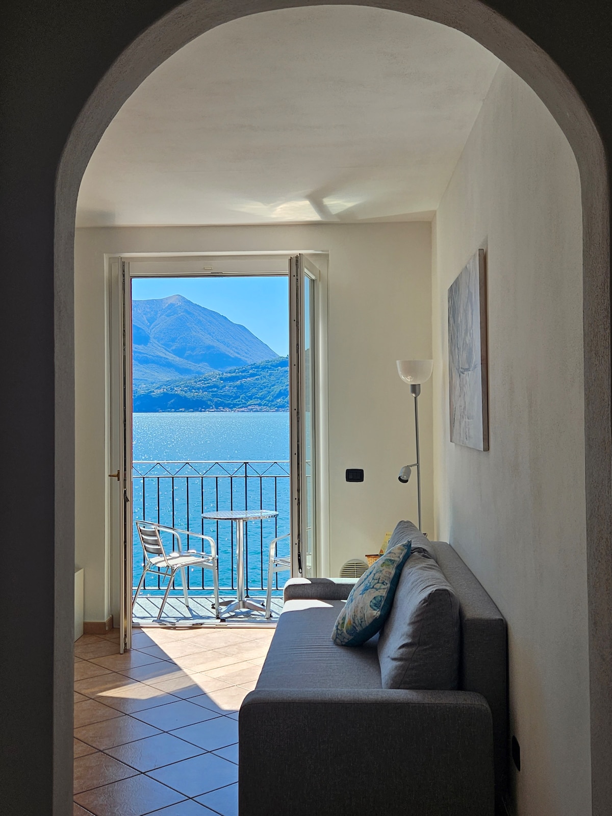 A cozy living space is framed by an archway, leading to a balcony overlooking the lake. A comfortable couch is positioned thoughtfully, while natural light enters through open doors. Scenic views of the mountains and water are visible from the space, enhancing the serene atmosphere.