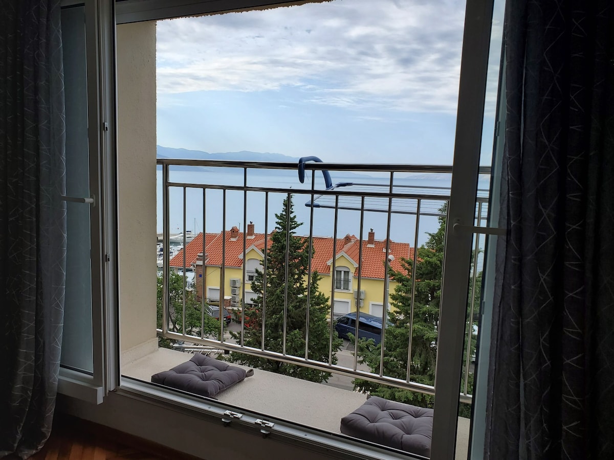 A French balcony provides a view of the surrounding landscape, featuring greenery and rooftops. The wide glass doors enhance natural light, and two cushions are placed on the balcony floor. Soft curtains frame the view, adding a gentle touch to the space.