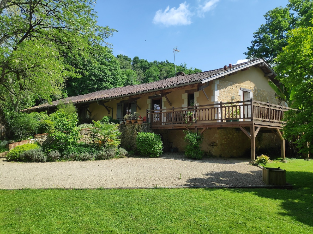 A spacious rural gîte is set against a backdrop of lush greenery. The building features a wooden terrace, adorned with potted plants, and overlooks a gravel pathway and a manicured lawn. Surrounding trees provide shade and enhance the tranquil setting.