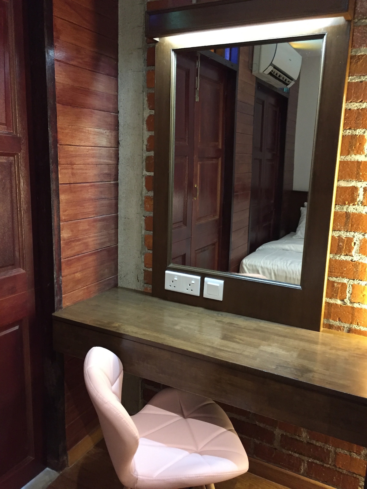 A compact vanity area features a wooden table and a pink upholstered chair. Above the table, a large mirror reflects the warm wooden tones of the surroundings. Electrical outlets are conveniently located near the mirror, providing easy access for personal devices.