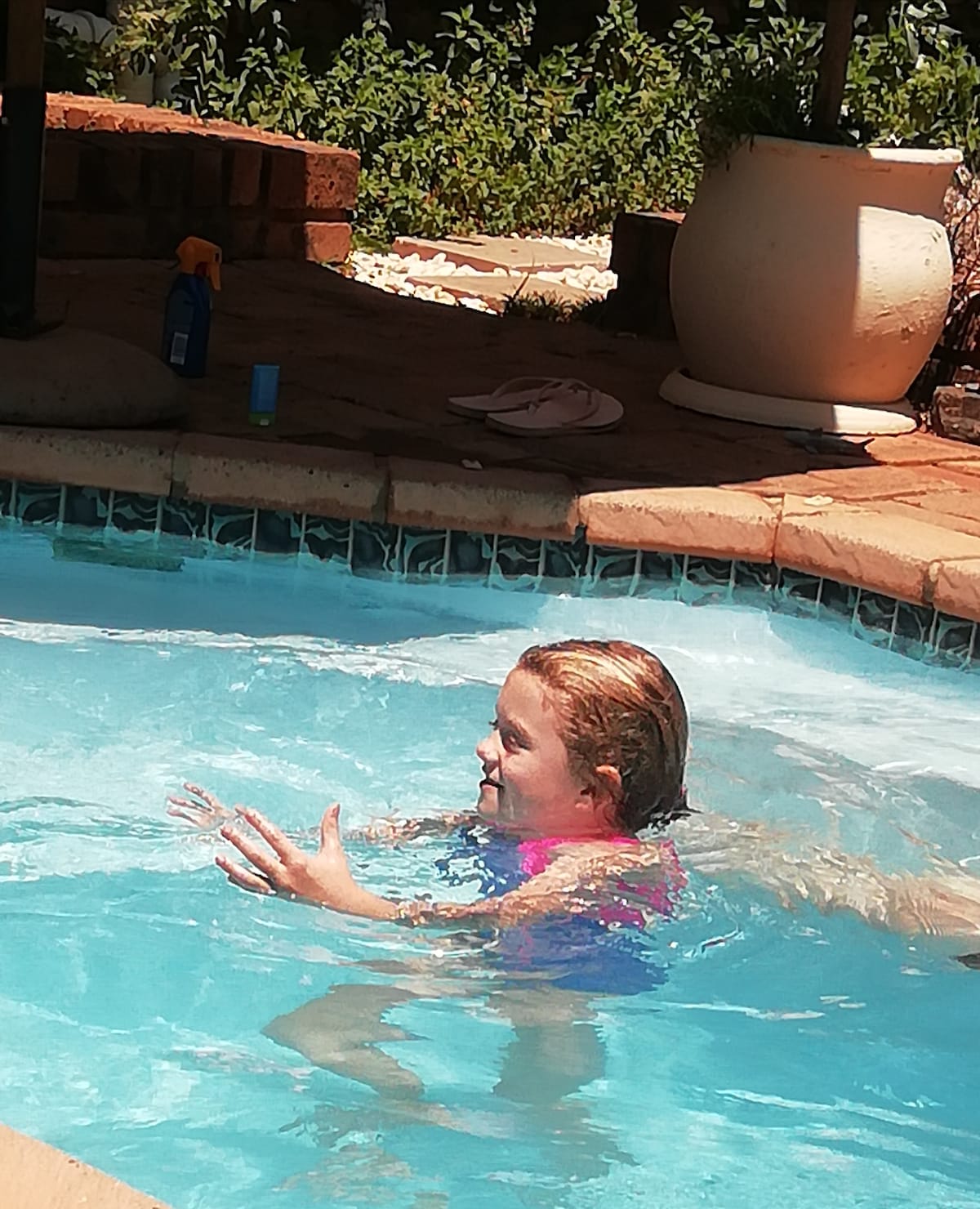 A splash pool area is visible, featuring clear blue water surrounded by a stone border. Sunlight reflects off the surface. A child is seen enjoying the water, with greenery and a terracotta planter in the background, contributing to a relaxed outdoor environment.