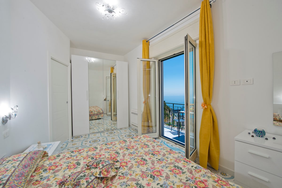 A bright bedroom features a spacious bed adorned with a floral-patterned duvet. Large glass doors open to a balcony, allowing natural light to fill the space. Vibrant yellow curtains frame the doors, while decorative mirrors and a bedside table enhance the airy atmosphere.