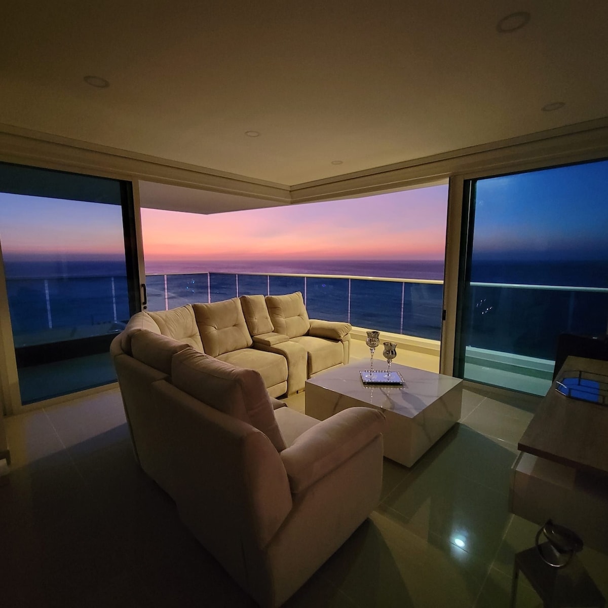A comfortable seating area is arranged with a light-colored sectional sofa facing expansive glass doors. A coffee table sits in the center, and the background features a stunning view of the ocean at sunset, with hues transitioning from orange to purple in the sky.