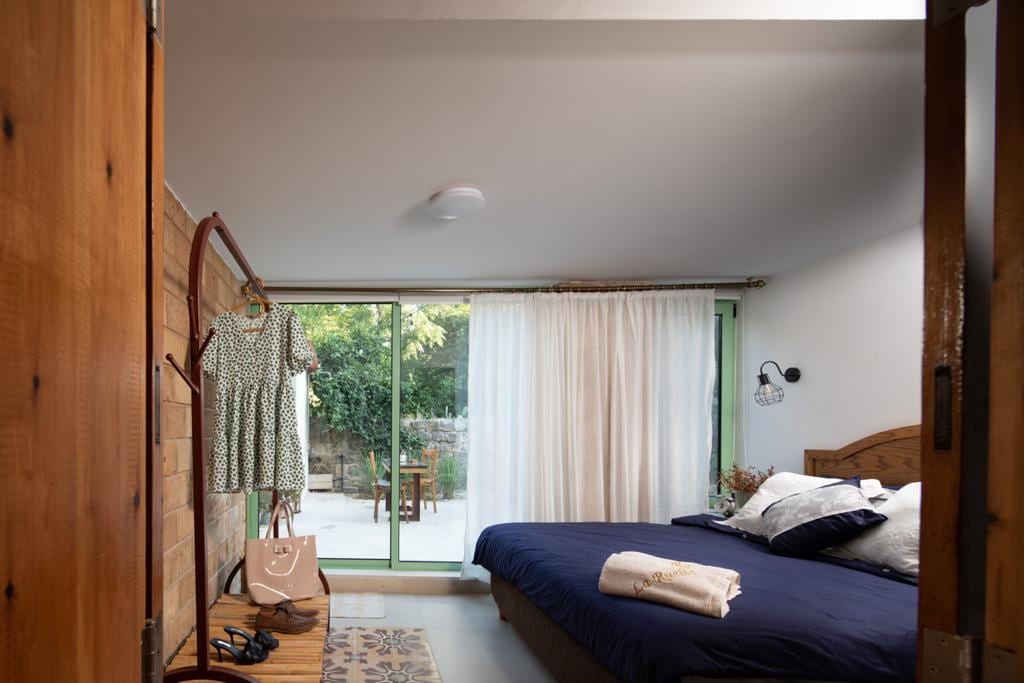 A bedroom is viewed through an open door, featuring a spacious bed with dark bedding. A wooden hanger displays a dress beside a bag. Natural light enters through a large glass door, leading to a garden space with outdoor seating.