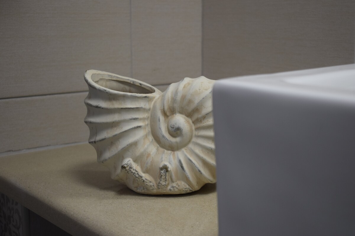 A decorative shell-shaped planter is positioned on a smooth, beige countertop. The planter displays a textured surface reminiscent of natural elements, while a modern white sink can be seen in the foreground, partially obscuring the view.