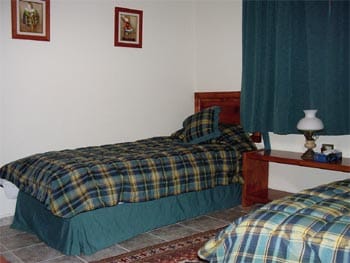 The sleeping area features two single beds with plaid bedding, complemented by coordinating bed skirts. A wooden nightstand holds a small lamp and clock. The room is softly lit, with green curtains framing a window, enhancing the cozy ambiance.