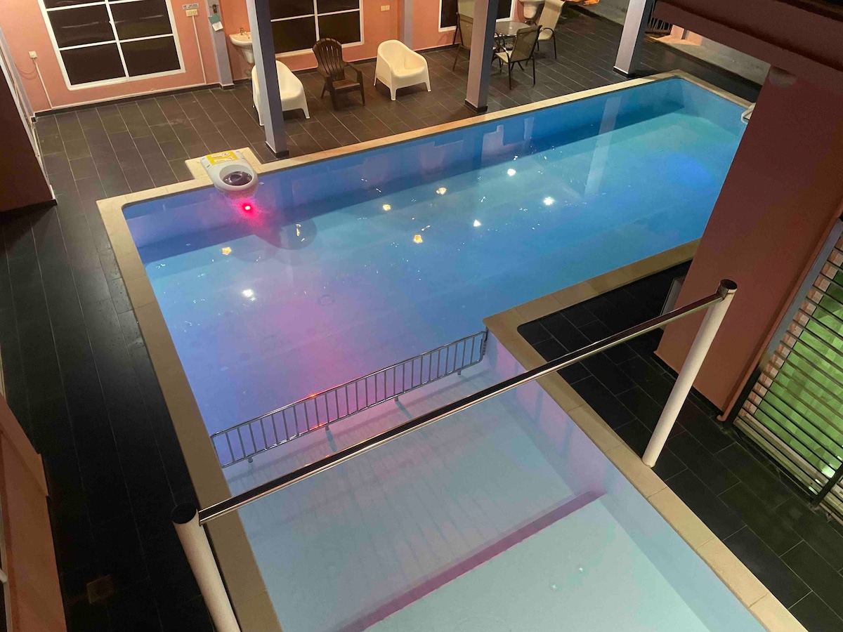 A spacious private pool is visible, featuring inviting turquoise water. Surrounding the pool are areas for seating, including white chairs. Soft lighting enhances the ambiance, while the layout allows for easy access to both indoor and outdoor spaces.