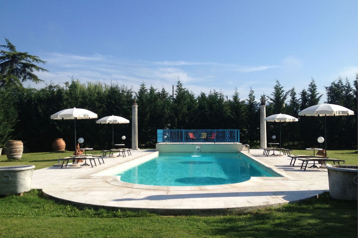 A sparkling swimming pool is framed by well-manicured grass and surrounded by elegant lounge chairs. Umbrellas provide shade for guests, while tall hedges offer privacy. The pool area is complemented by decorative planters, creating a serene outdoor retreat.