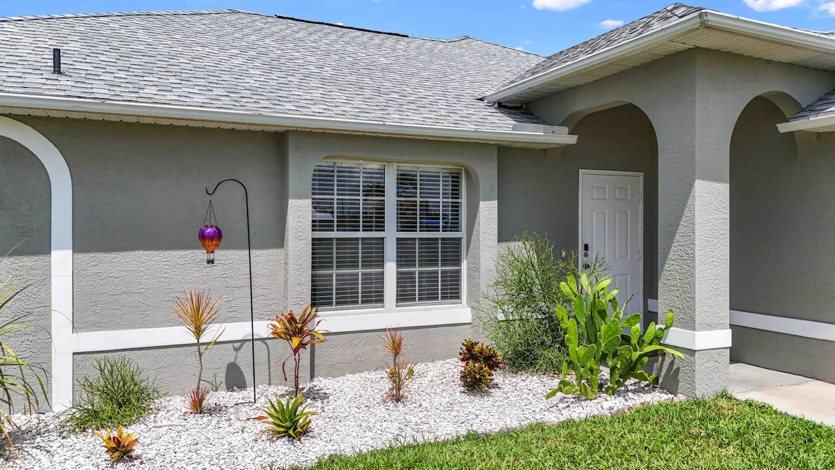 The exterior of the home features a well-maintained front garden with various plants and vibrant accents. A large window allows natural light into the interior, while a welcoming entrance door is positioned to the right. The overall appearance reflects a clean and inviting aesthetic.
