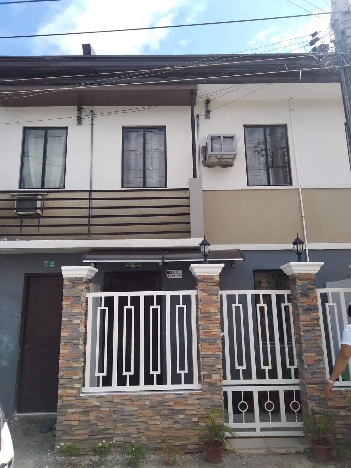 The exterior of a two-story house is displayed, featuring a mixture of white and beige walls. A balcony is visible above the entrance, and a well-maintained stone fence delineates the property. A small garden area is present at the base of the fence.