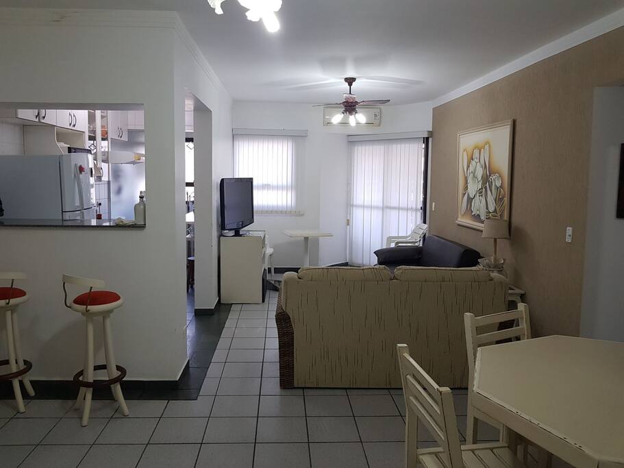 Entrada do apartamento com cozinha americana seguida da sala de Tv e sala de jantar com saida para sacada com vista para o mar e area de lazer e ar condicionado.