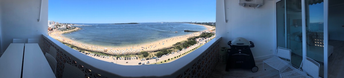 A panoramic view captures the expansive coastline, showcasing golden sands and the shimmering water. The scene includes a spacious balcony area with a grill, providing an ideal outdoor setting for relaxation. Clear blue skies enhance the overall brightness of the landscape.