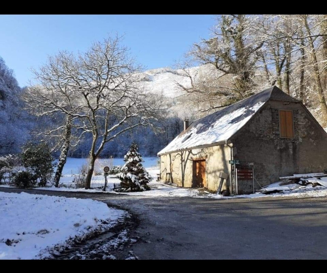 A rustic barn is surrounded by a serene winter landscape, blanketed in snow. Snow-covered trees frame the scene, while distant mountains rise under a clear blue sky. The barn's weathered exterior and orange shutters contrast gently with the white surroundings.