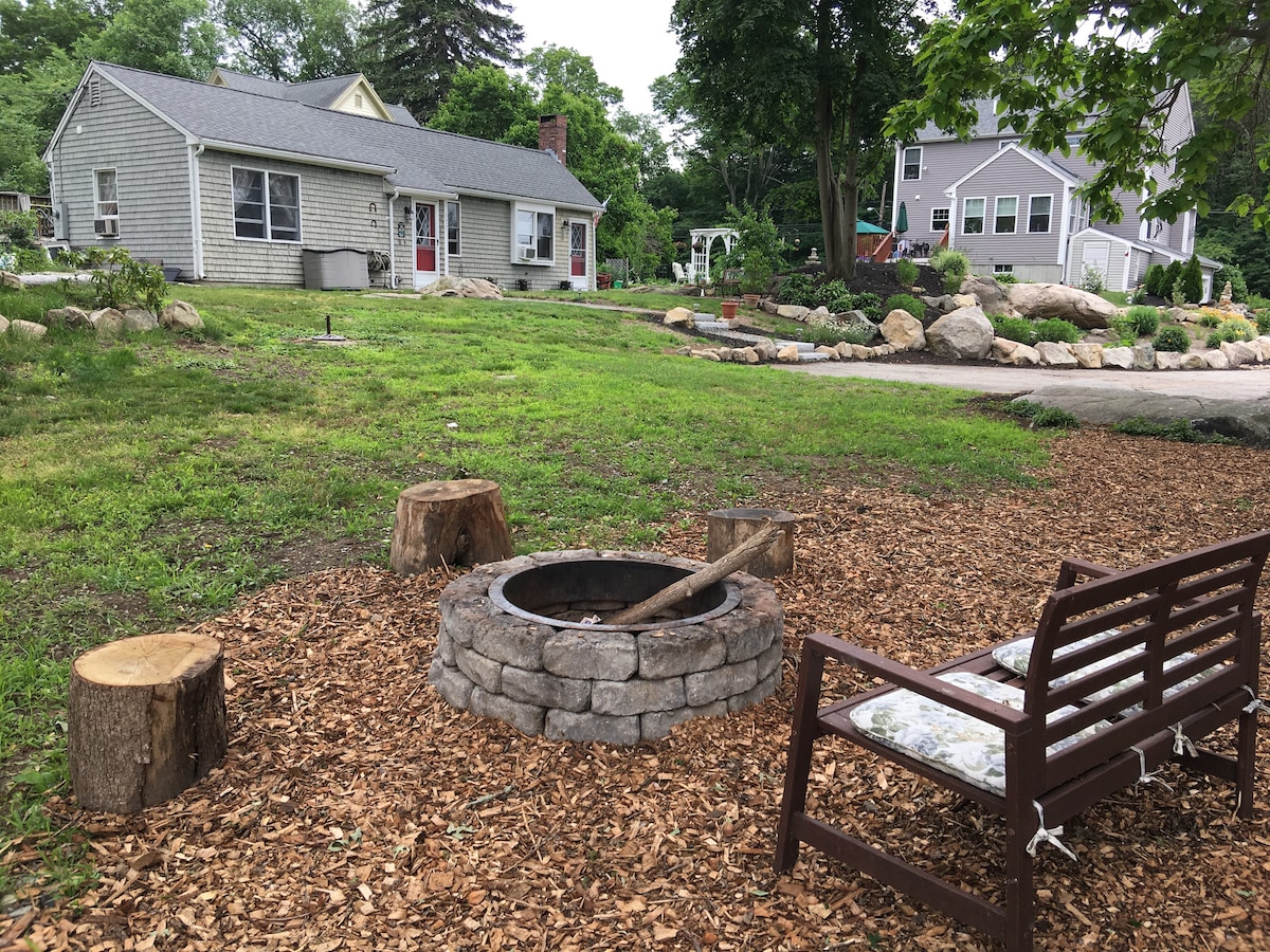 A fire pit made of stone is surrounded by wooden seating and mulch, providing a cozy outdoor gathering space. A grassy area leads to a cottage with a gray exterior and a sloped roof, framed by trees and neighboring homes in the background.