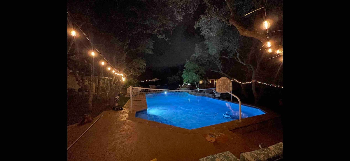 A well-lit pool glows in blue tones under the night sky, surrounded by trees. Decorative string lights drape overhead, enhancing the outdoor ambiance. A volleyball net is positioned at one end of the pool, contributing to the recreational setting.