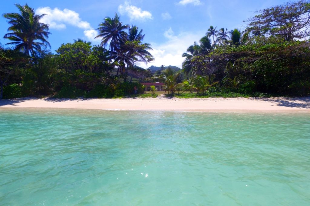 The image captures a serene beach scene with soft, white sand and clear turquoise waters. Lush palm trees and tropical vegetation frame the landscape, providing a backdrop of greenery. The tranquil lagoon reflects the vibrant sky, highlighting the peaceful setting of the property.