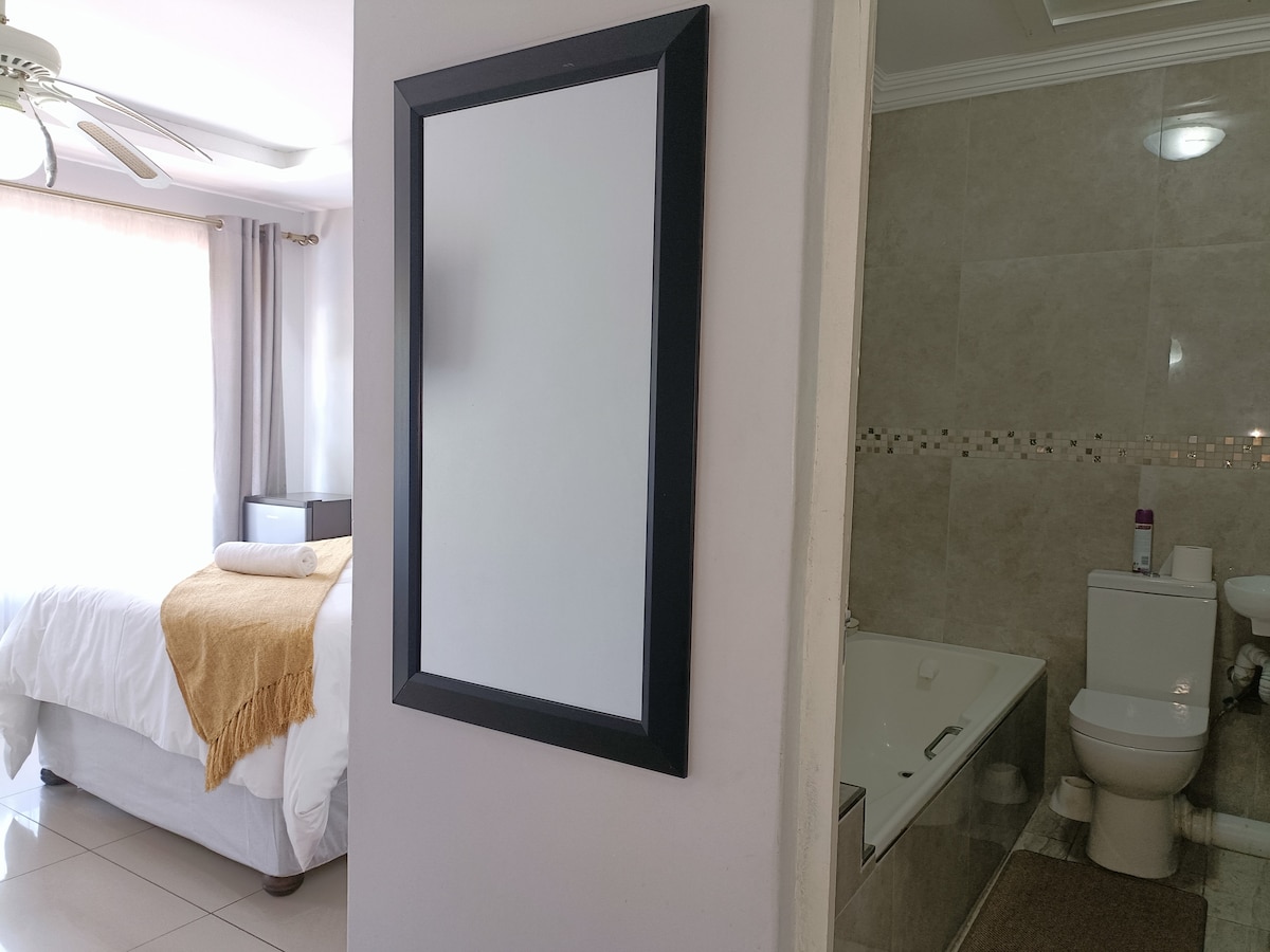 A wall-mounted mirror is prominently displayed next to an en-suite bathroom. The spacious bedroom can be seen in the background, featuring a bright bed with a light-colored throw. The bathroom includes a bathtub and toilet, with neutral tiles and subtle decorative elements.