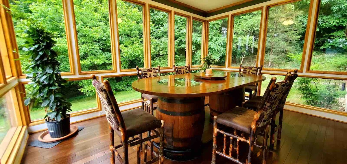 A dining room is presented with large windows offering views of surrounding greenery. A round table crafted from wood, resembling a barrel, is surrounded by six wooden chairs. A potted plant is positioned near the window, enhancing the natural ambiance of the space.