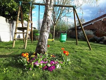 A garden area is depicted featuring a swingset with two swings hanging from a sturdy frame. Vibrant flowers, including tulips and pansies, bloom at the base of a tree, surrounded by lush green grass under a clear sky.