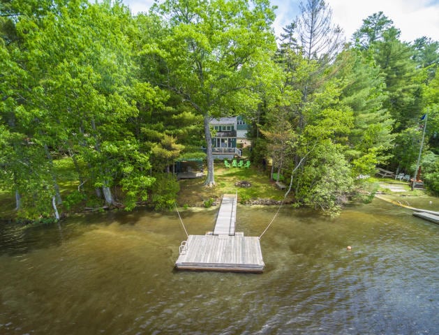 A wooden dock extends into the calm waters, framed by lush greenery. A cottage can be seen in the background, surrounded by trees. Several white chairs are positioned on the lawn, providing a space to relax and enjoy the water views.