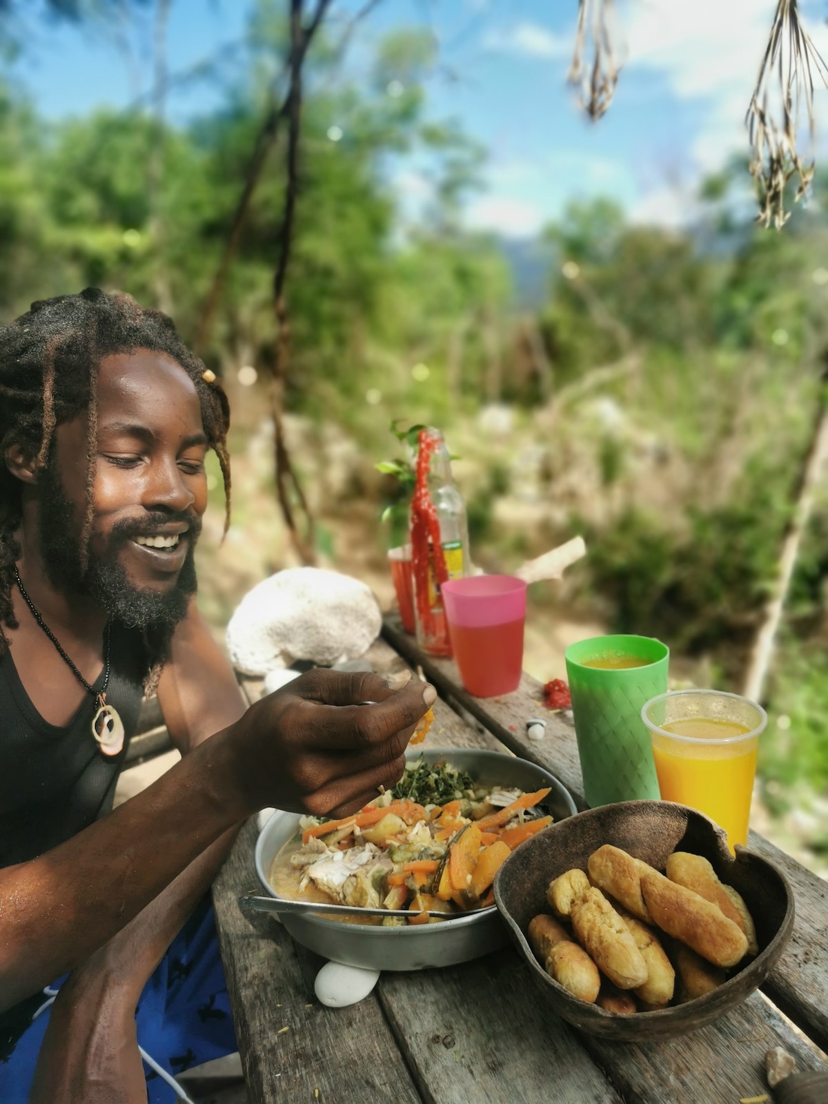 A wooden table is set outdoors, displaying a vibrant meal featuring a variety of vegetables, rice, and dumplings. Colorful drinks are placed in cups nearby, surrounded by lush greenery and a distant landscape. A hand is shown holding a fork, bringing food to the mouth.