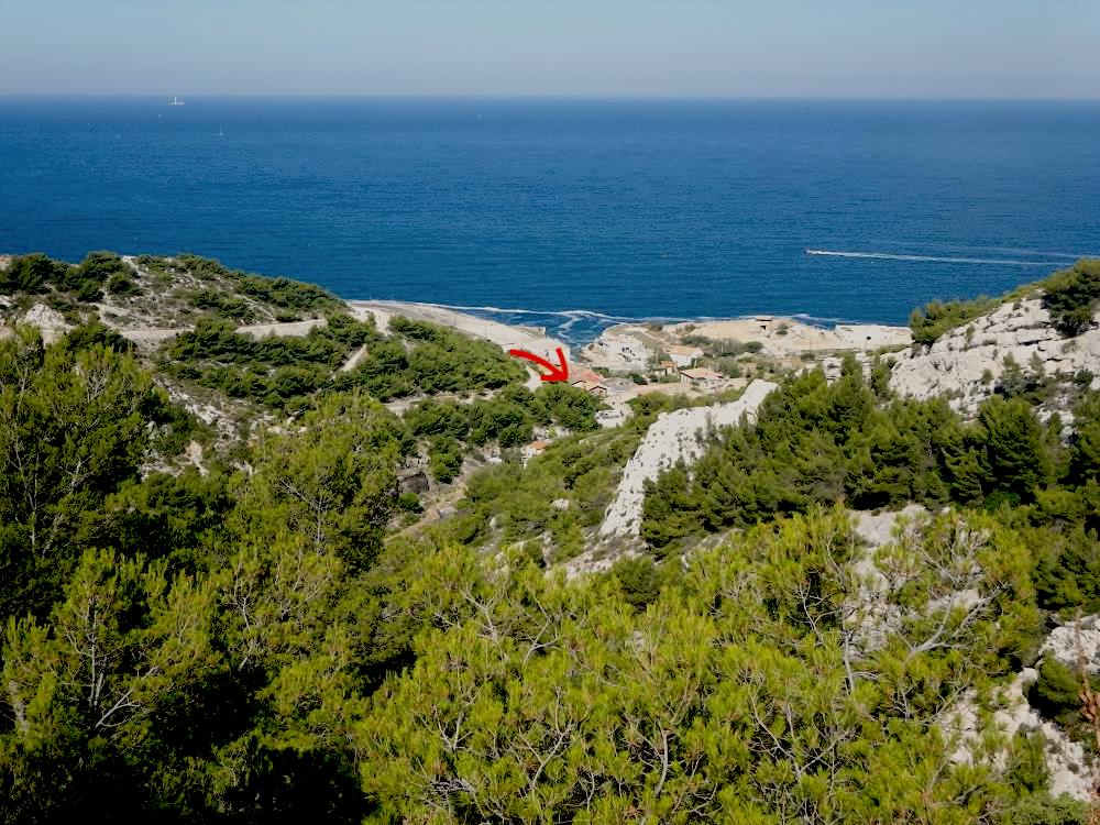 A serene coastal view captures the expansive blue sea under a clear sky, with rocky cliffs and greenery in the foreground. The shoreline, featuring a mix of sand and pebbles, outlines the landscape, inviting exploration and connection with nature.