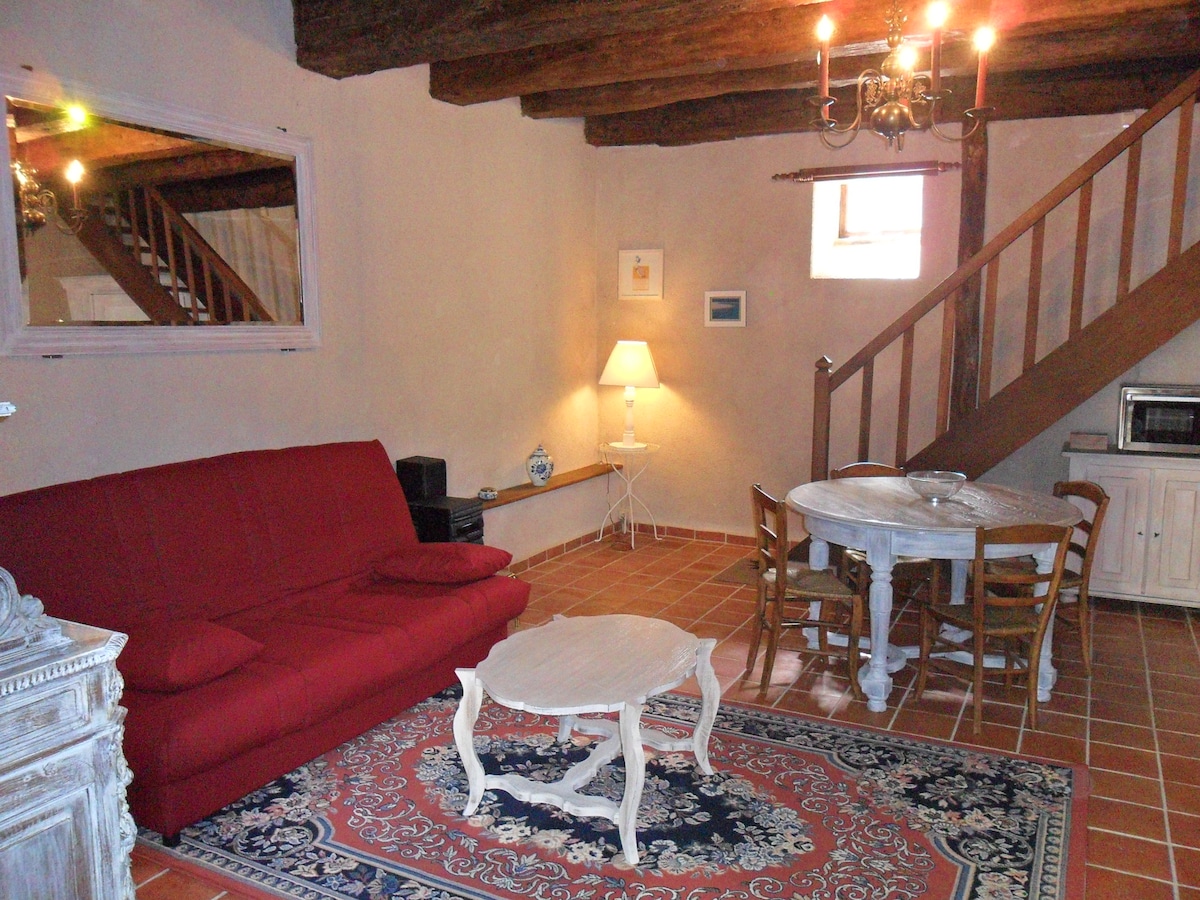 A living area is visible, featuring a red sofa and a small white coffee table on a patterned rug. A dining table with four wooden chairs is set against the wall, with a staircase leading to an upper floor. Natural light enters through a nearby window.