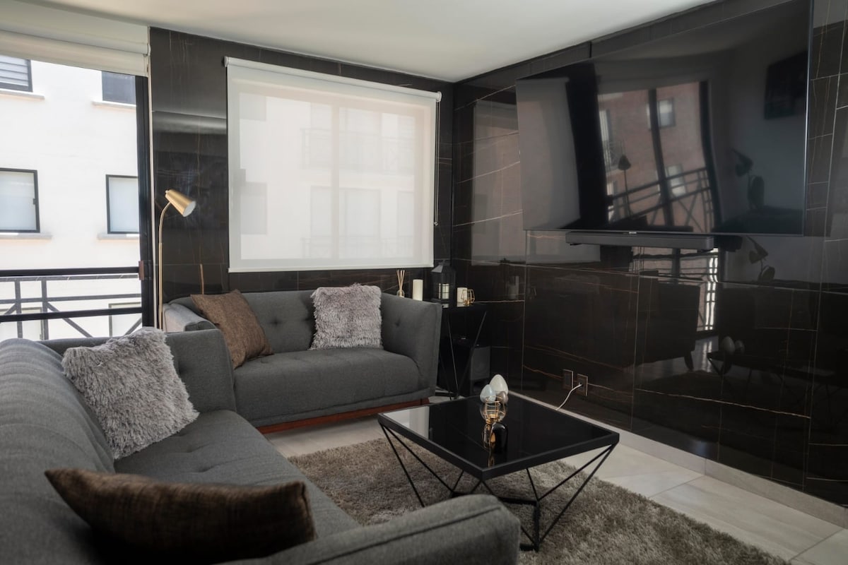 A modern living area features a gray sectional sofa adorned with plush throw pillows. A sleek black coffee table sits at the center, with a decorative lamp illuminating the space. Large windows allow natural light to fill the room, reflecting off dark, glossy walls.