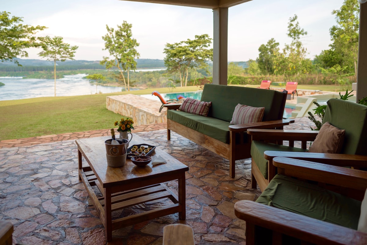 A shaded outdoor seating area features wooden furniture with green cushions, complemented by a rustic coffee table. Fresh flowers in a vase and a bowl of fruit are displayed on the table. The nearby infinity pool offers views of the expansive river and surrounding greenery.