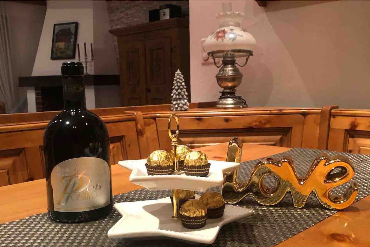 A cozy dining table is displayed with a two-tiered serving stand holding chocolate treats, alongside a decorative golden 'love' sign. A bottle of wine is positioned prominently, with an elegant lamp and a small Christmas tree in the background, adding to the inviting atmosphere.