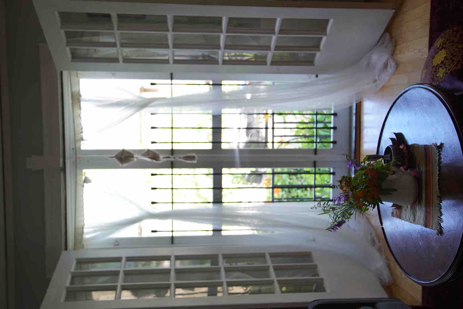Large glass doors allow natural light to flood the room, framed by sheer curtains that gently sway in the breeze. A round coffee table is positioned in front, surrounded by a potted plant that adds a touch of greenery. The view outside displays a lush garden area.