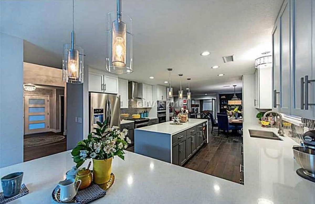 The kitchen area presents a modern design with an expansive island featuring a white countertop. Pendant lights hang above, illuminating the space. Stainless steel appliances are visible in the background, alongside a dining area with dark wood furniture. A touch of greenery is added by a flower arrangement.