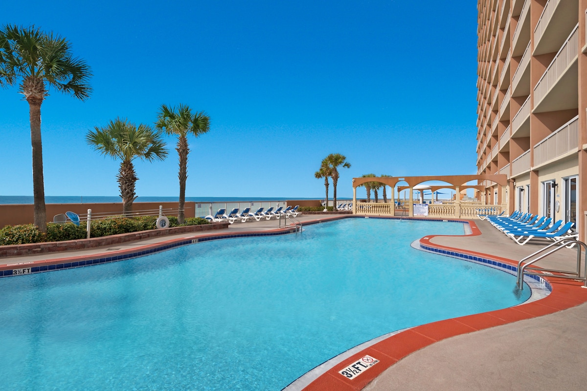 A relaxing pool area is surrounded by palm trees, featuring a clear blue pool with a gentle curved edge. Lounge chairs are positioned around the pool, facing the ocean, while a white gazebo provides shaded seating. The bright blue sky enhances the tranquil ambiance.