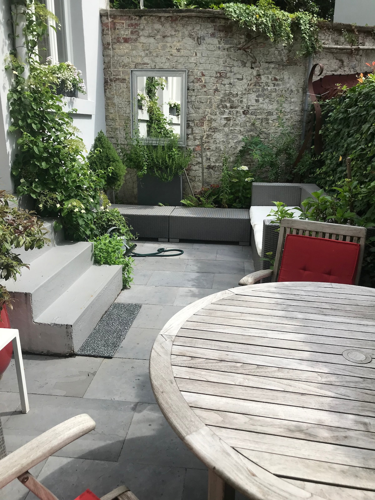 An outdoor terrace area features a circular wooden table surrounded by several chairs. Lush greenery is present, including climbing plants against the walls. A small set of stairs leads up to the entrance, and a mirror reflects the natural surroundings.