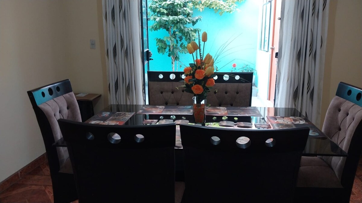 A dining area is presented with a glass table surrounded by four upholstered chairs, each featuring a unique circular design. A vibrant flower arrangement sits at the center of the table, while large windows allow natural light to brighten the space.