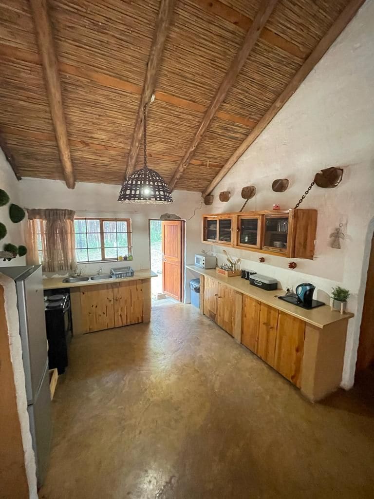 The kitchen area features rustic wooden cabinetry and appliances, with a large window allowing natural light to fill the space. A woven light fixture hangs above, complementing the thatched roof. Nearby, the entrance door opens to the outside, enhancing accessibility.
