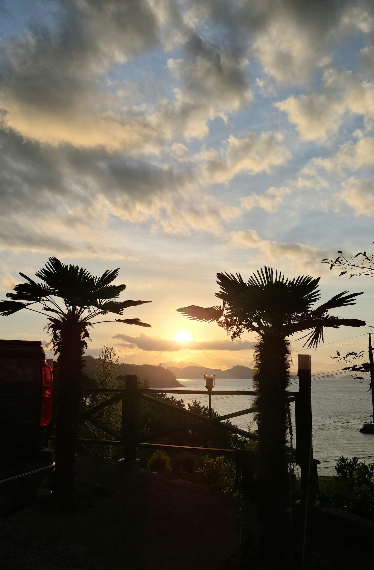 Silhouettes of palm trees frame a serene sunset over the horizon, with subtle reflections on the calm water. Soft clouds are illuminated by golden hues, adding to the tranquil ambiance of the coastal setting.