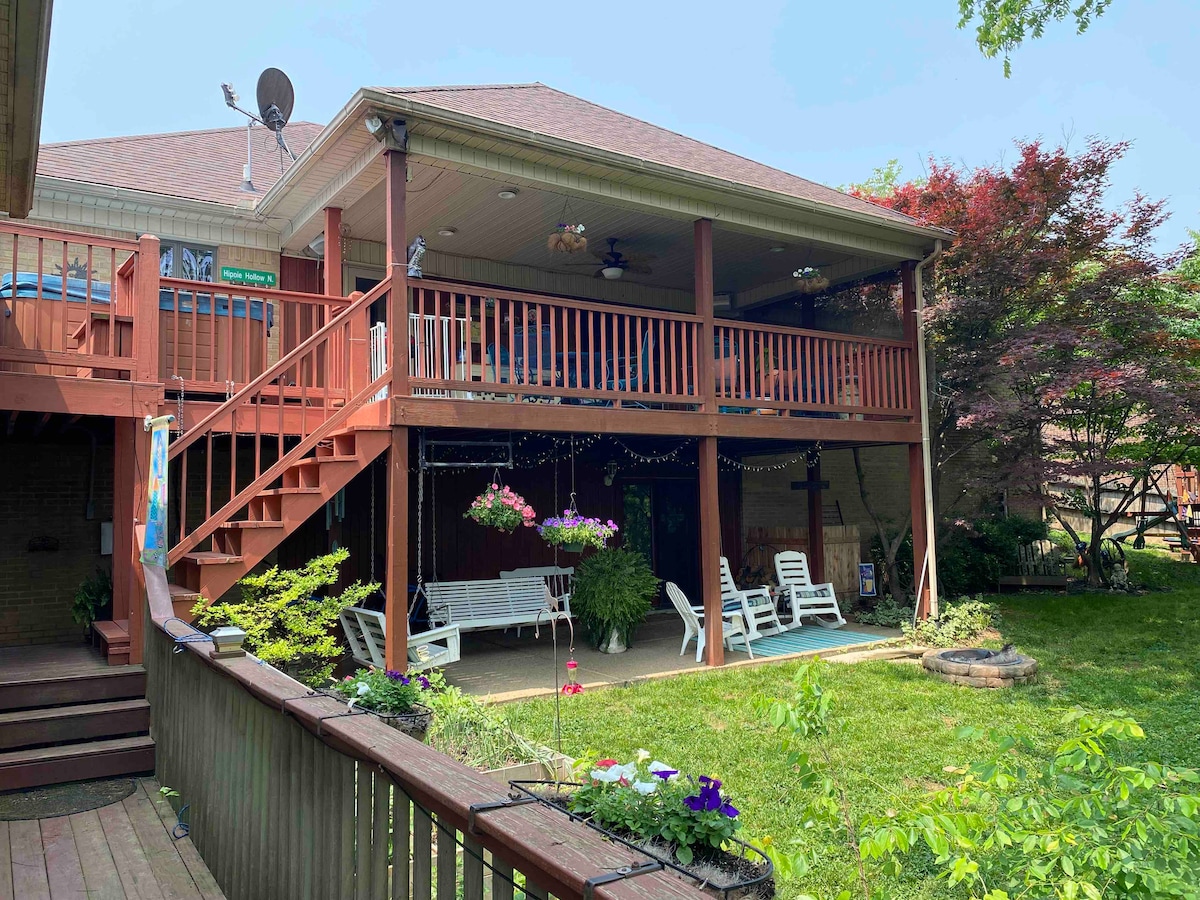 The outdoor space features a large porch with ceiling fans and multiple swings. Beneath the porch, a well-maintained yard is surrounded by greenery, with flowering plants adding color. A small stone feature is visible on the ground, enhancing the tranquil atmosphere.