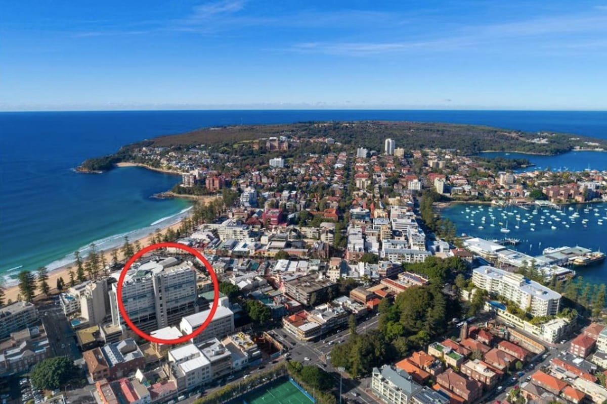 An aerial view captures a coastal area in Manly, highlighting the proximity of the apartment to the beach and nearby amenities. The layout of the surrounding buildings and the ocean view creates a sense of location, with vibrant landscapes encompassing the area.
