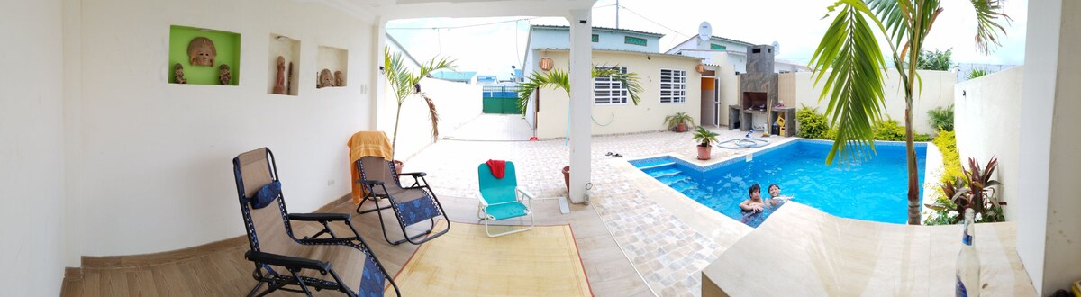 An outdoor area features a spacious pool surrounded by tropical plants and lounge chairs. A patio with a light-colored rug is visible, alongside a shaded seating space. The house in the background showcases large windows, contributing to the open and inviting atmosphere.
