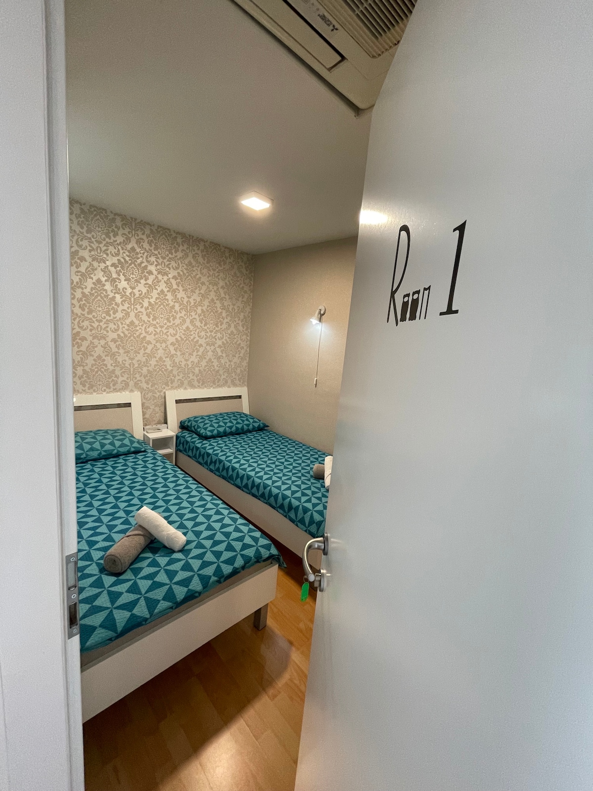 A cozy room is displayed with two neatly made beds featuring patterned blue bedding. The walls are adorned with a subtle decorative design. A small lamp is mounted beside a bed, and light is provided by a ceiling fixture. A door is visible leading to the hallway.