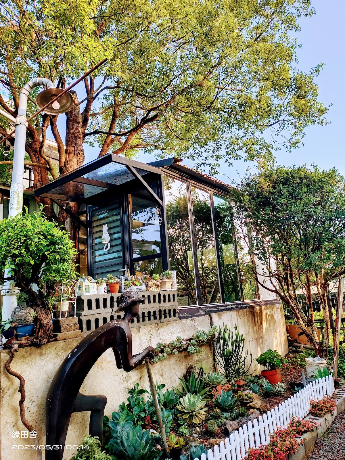 A charming outdoor space is highlighted by a glass structure surrounded by lush greenery and a variety of potted plants. A small, white picket fence encloses the landscaped area, adding a quaint touch to the setting.