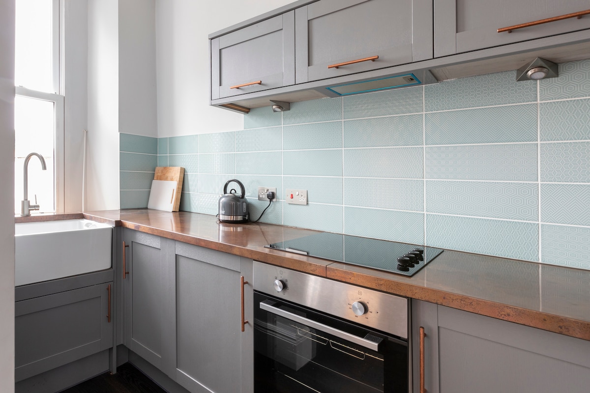 The kitchen features modern cabinetry in a soft grey tone, complemented by a mint green tiled backsplash. A stainless steel oven and hob are positioned alongside a sink. A kettle and wooden cutting board add functional details to the space.