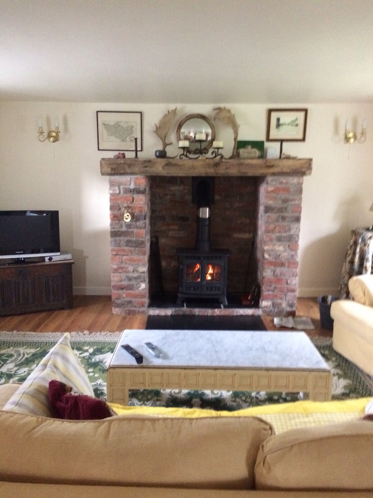 A cozy sitting area features a brick fireplace with a wood-burning stove, providing warmth and comfort. A television is positioned on a dark wooden unit opposite a light-colored sofa, complemented by decorative pillows. Framed artwork is displayed on the walls, enhancing the cottage's character.