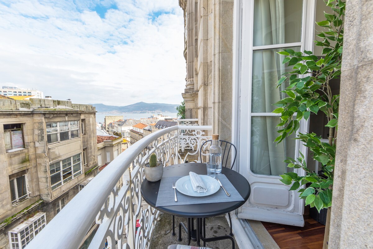 A small balcony is featured with a round black table set for two, including a glass of water and neatly folded napkins. Lush greenery is visible nearby, enhancing the view of the cityscape and distant water. The ornate railing adds architectural interest.