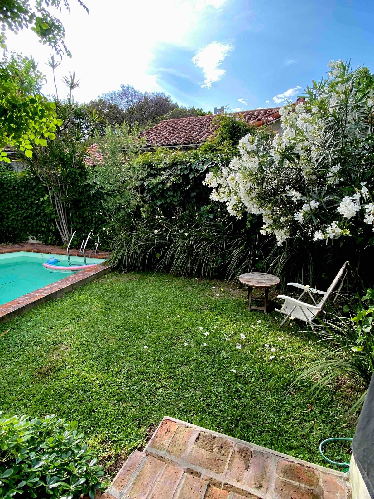 A serene garden area features a small pool surrounded by lush greenery and flowering plants. A rustic table and a white chair sit on the grass, creating a tranquil space for relaxation. The brick pathway leads to the refreshing pool area under a clear blue sky.